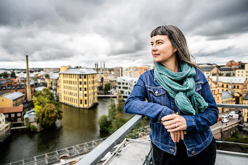 A photo of Miriah smiling with a Swedish urban landscape in the background. She has
  long hair and short bangs.